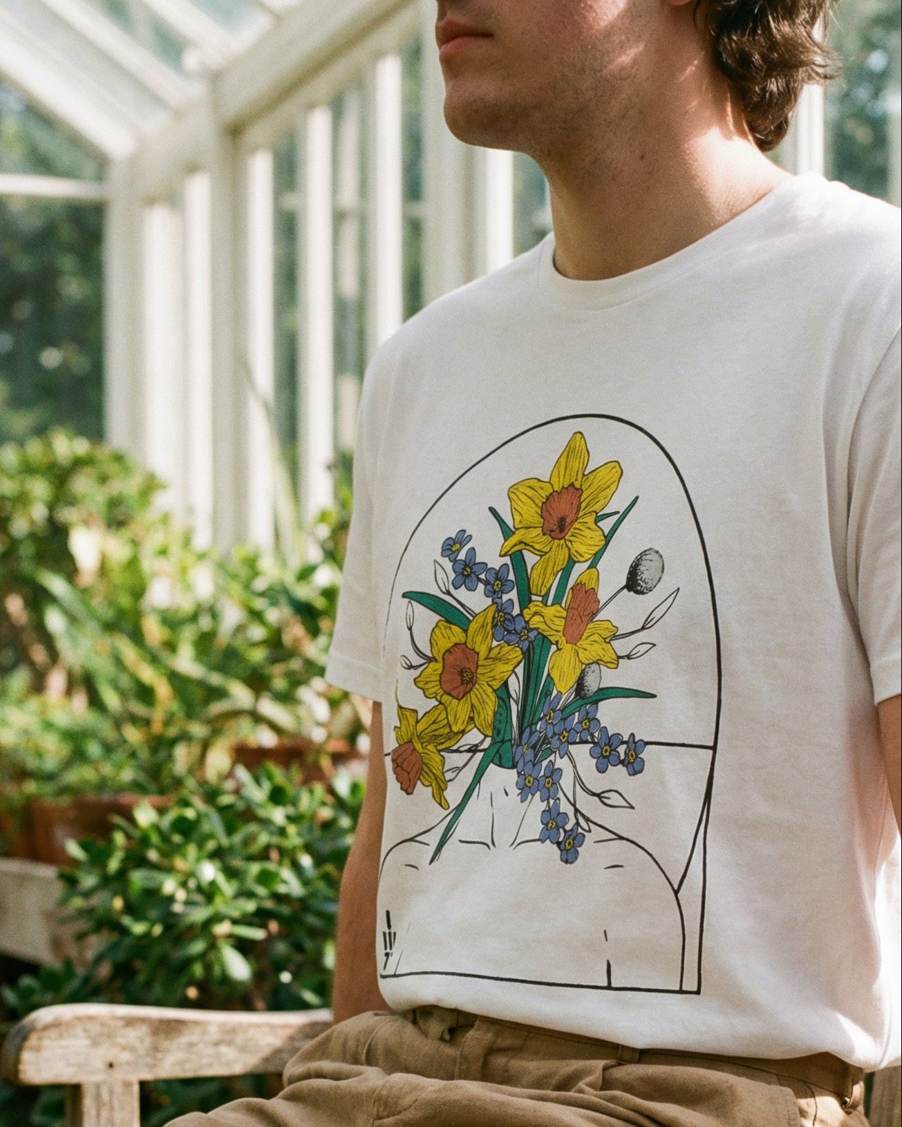 Person wearing a white t-shirt with a floral design sitting in a greenhouse.
