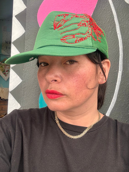 Person wearing a green cap with red lobster embroidery against a colourful wall.
