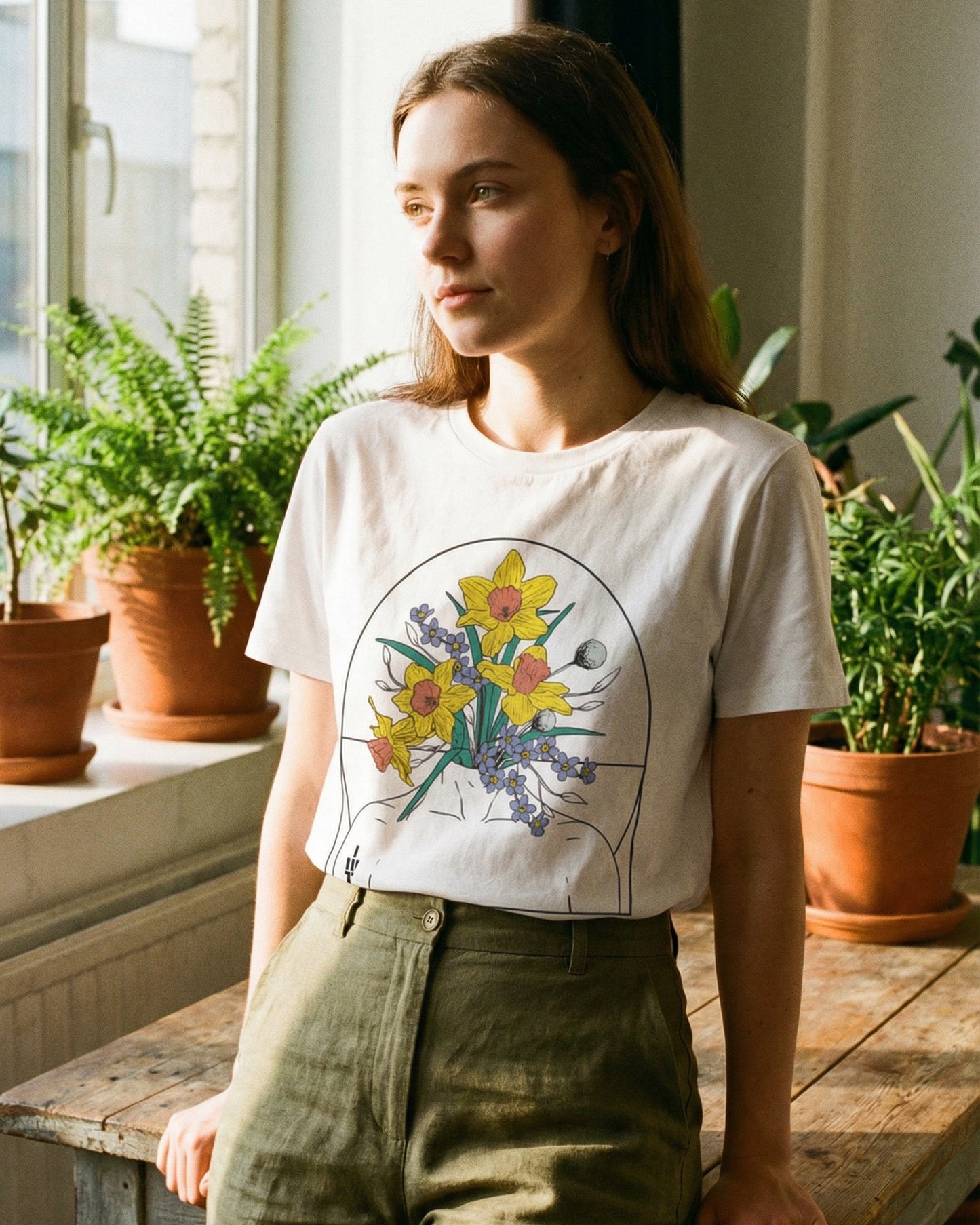 A woman wearing a white t-shirt with a daffodil and forget-me-not design in a room with plants in terracotta pots