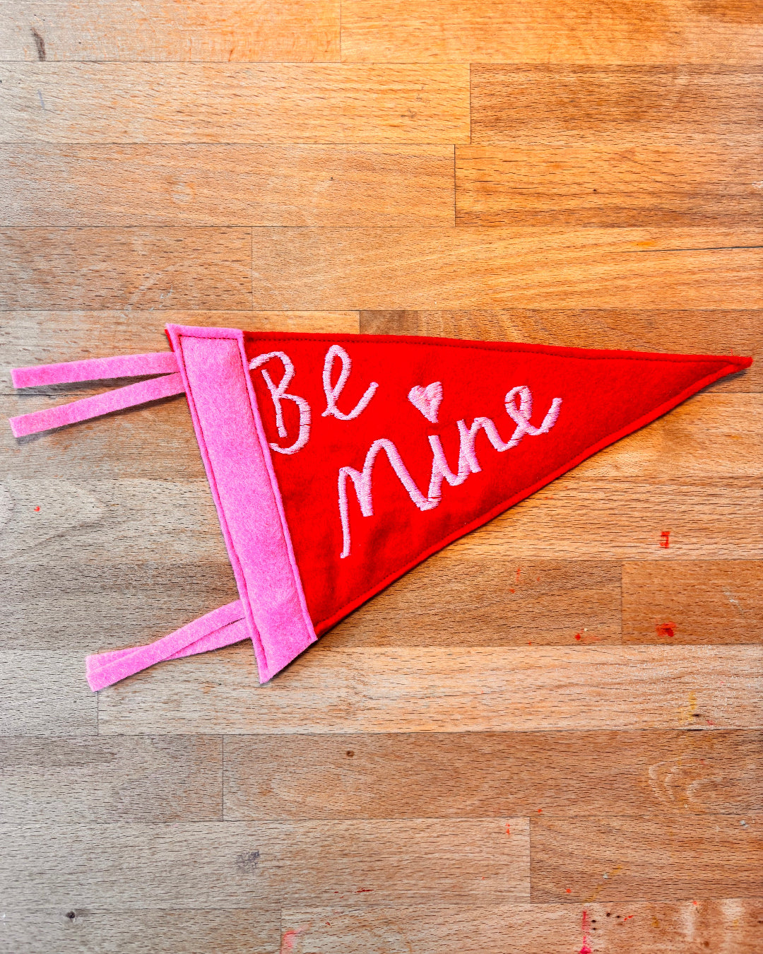 Red and pink fabric banner with 'Be Mine' text on a wooden surface