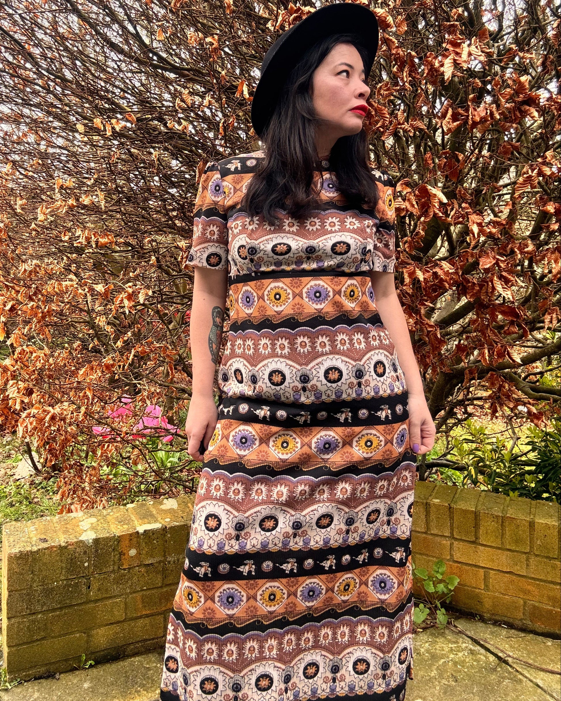 Person wearing a patterned 1970s vintage boho dress standing outdoors with trees and grass in the background.