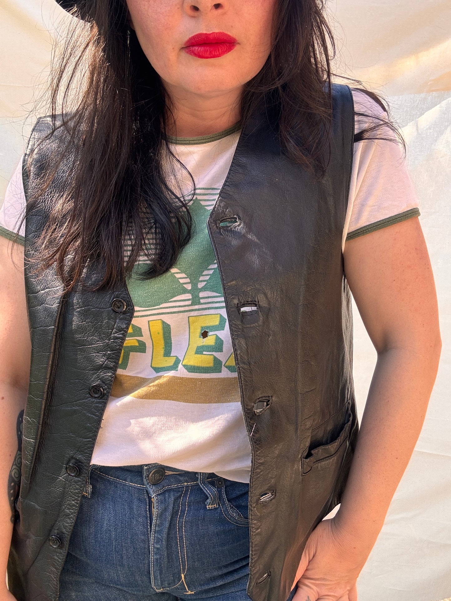 Person wearing a black leather vest over a white shirt with green text and blue jeans.