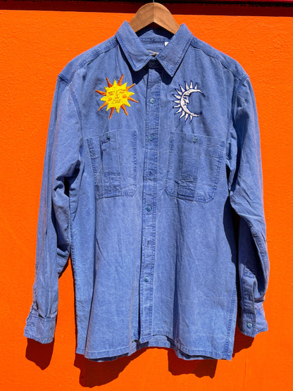 A blue denim button-up shirt hanging on a wooden hanger against a bright orange wall, featuring a yellow embroidered sun on the left chest and a silver embroidered crescent moon on the right chest.