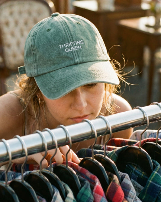 Person wearing a 'Thriftling Queen' cap looking at clothes on a rack.