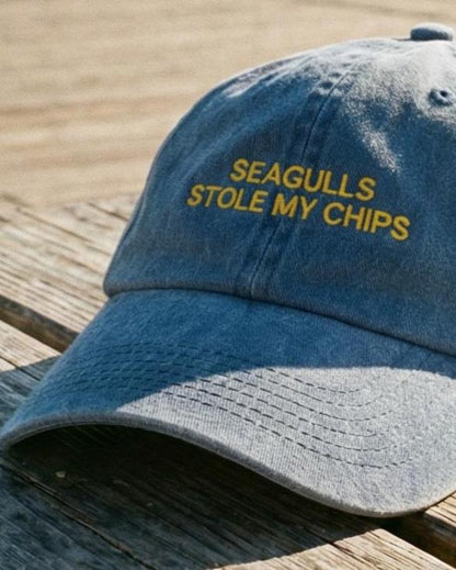 Blue denim cap with 'SEAGULLS STOLE MY CHIPS' text on a wooden surface