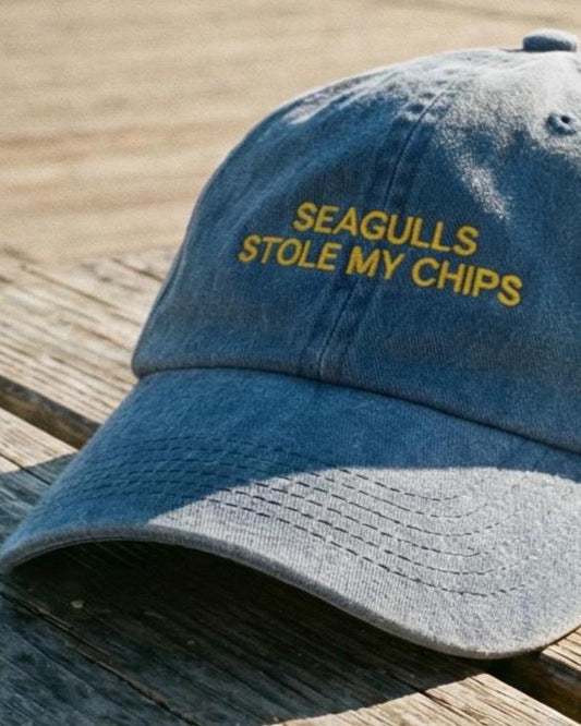 Blue denim cap with 'SEAGULLS STOLE MY CHIPS' text on a wooden surface